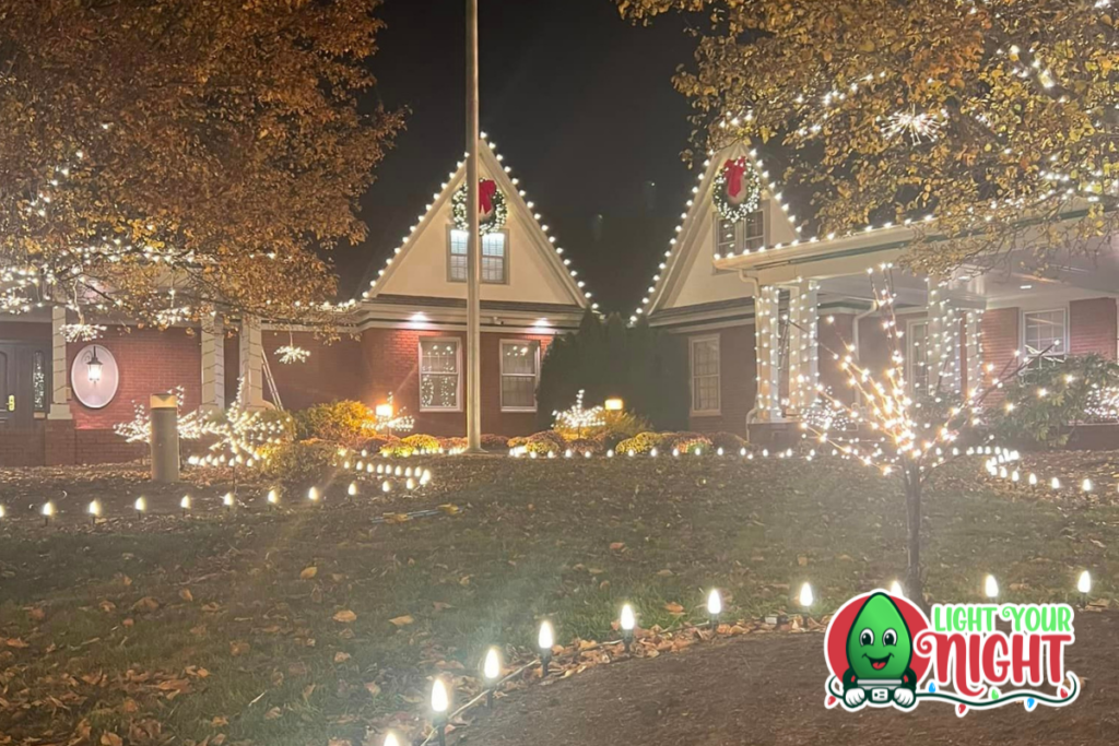 A festive house decorated for the holidays at night, featuring strings of white lights around the roof, columns, and trees. The front yard is outlined with lights. The "Light Your Night" logo featuring a smiling holiday bulb is in the bottom right corner, proudly cleaned by SUDS Power Washing.