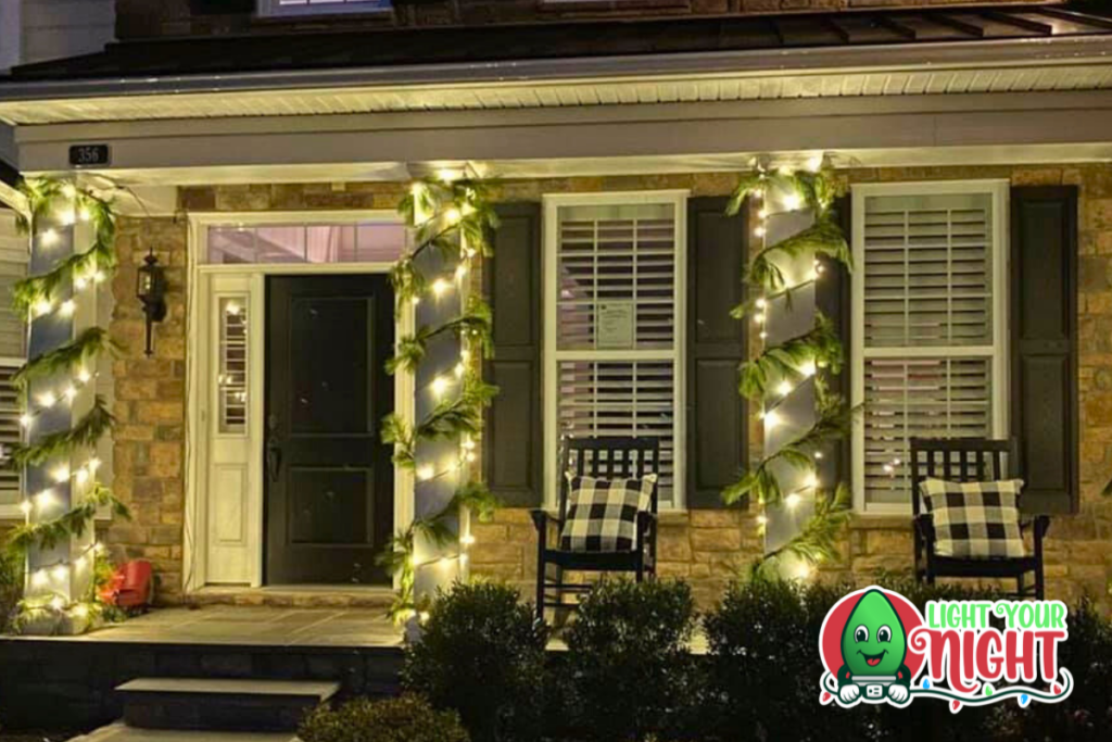 A house entrance decorated with festive lights and greenery wrapped around the pillars and railings. Two black rocking chairs with plaid cushions sit on the porch, freshly cleaned by SUDS Power Washing. A "Light Your Night" logo with a green character is at the bottom right corner of the image.