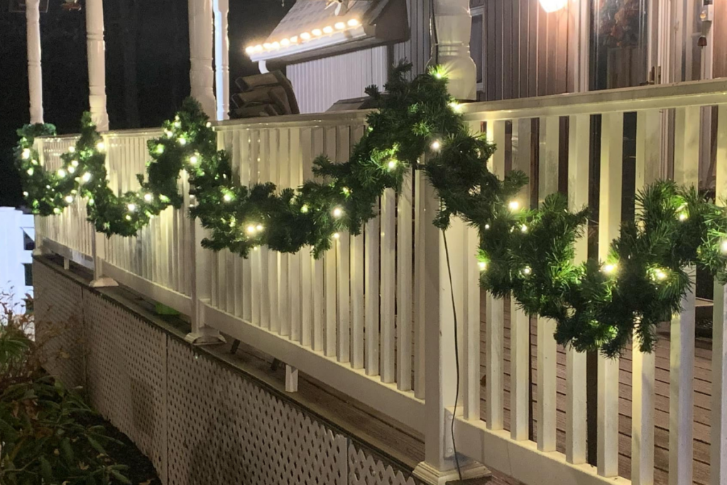 A porch is decorated with a garland of greenery wrapped around the white railing, illuminated by small white lights. The setup is part of a festive display, likely for the holiday season. The porch light is on, adding to the warm ambiance of the scene.