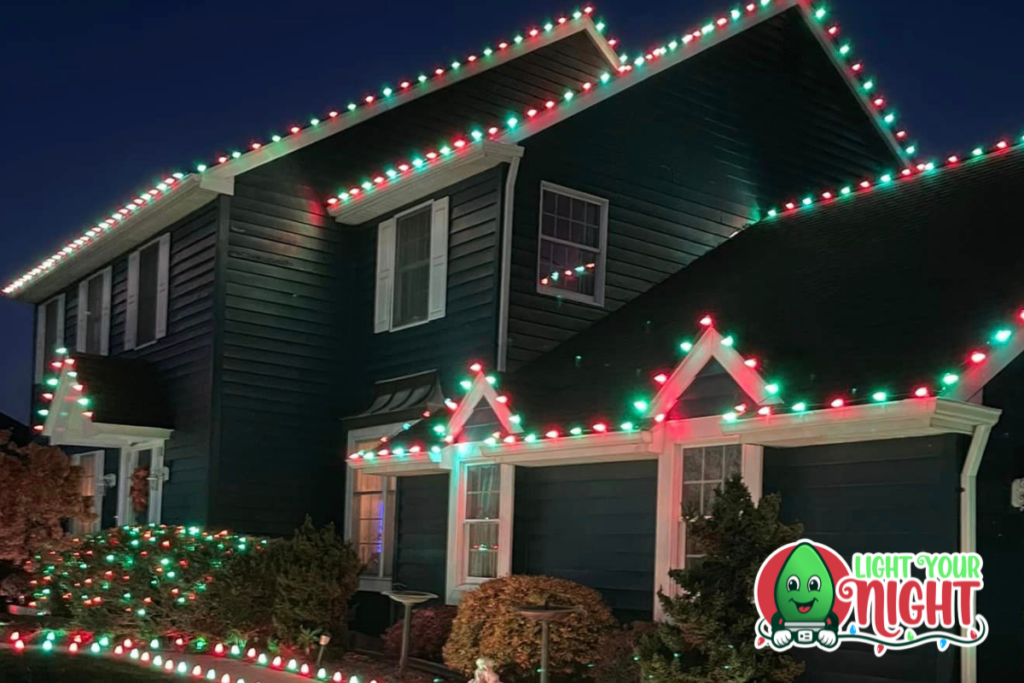 A large, two-story house is adorned with festive Christmas lights in red and green, creating a cheerful holiday display. The lights outline the roof and windows, and a "Light Your Night" logo with a smiling bulb character is seen in the bottom-right corner.