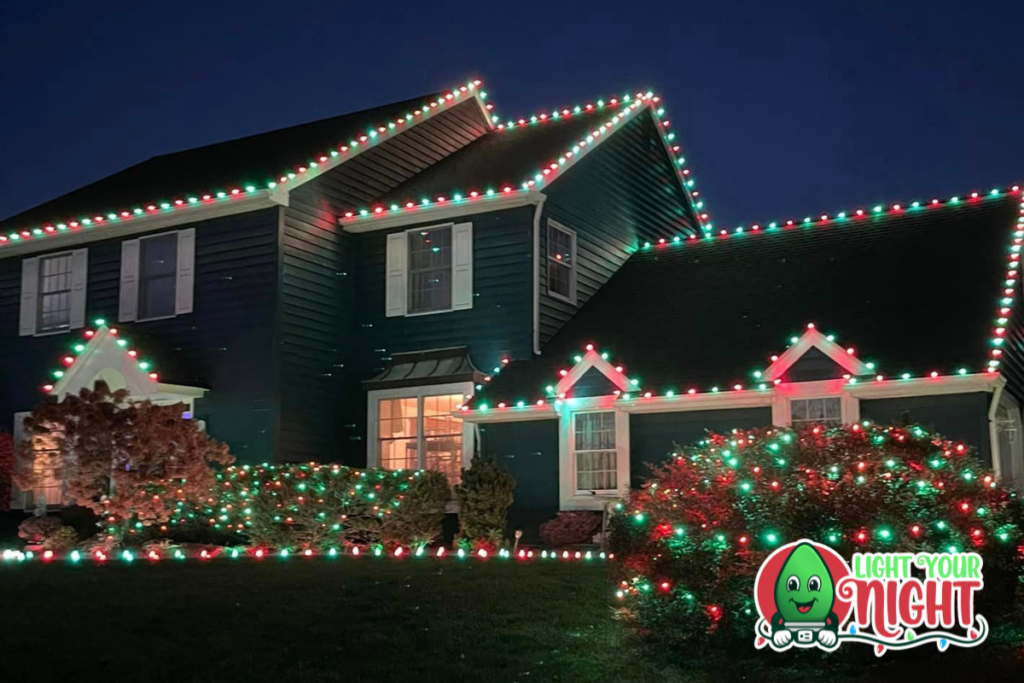 A house is decorated with festive Christmas lights, including red, green, and white lights outlining the roof and windows. Bushes around the house are also covered in colorful lights. A "Light Your Night" logo featuring a smiling light bulb character is in the bottom right corner.