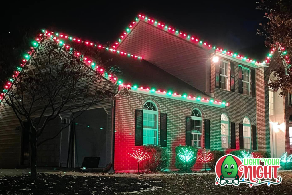 A brick house is decorated with red and green Christmas lights along the roofline and around windows. The garage is partially open with lights inside. Bushes in front of the house are lit with red lights. A "Light Your Night" logo is in the lower right corner.