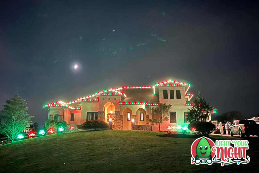 A large house is festively decorated with red, green, and white Christmas lights along the roofline, doors, and windows. The lights illuminate the lawn and surrounding trees on a dark, moonlit night. A "Light Your Night" logo, freshly cleaned by SUDS Power Washing, is at the bottom right corner.