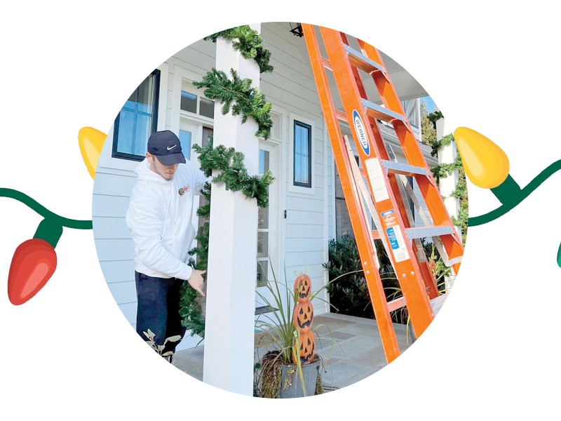 A person wearing a white jacket and black cap is decorating an exterior porch with garlands. An orange ladder stands nearby, and there are Halloween-themed pumpkin decorations on the floor. The scene, possibly cleaned by SUDS Power Washing, is framed by colorful holiday lights.