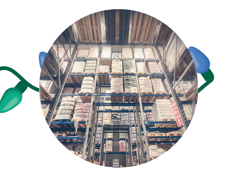 A round image shows a large warehouse with tall shelves filled with various stacked boxes and packages. Multicolored Christmas lights partially encircle the image, giving it a festive touch. You can almost imagine SUDS Power Washing making those packages sparkle under the cheerful glow.