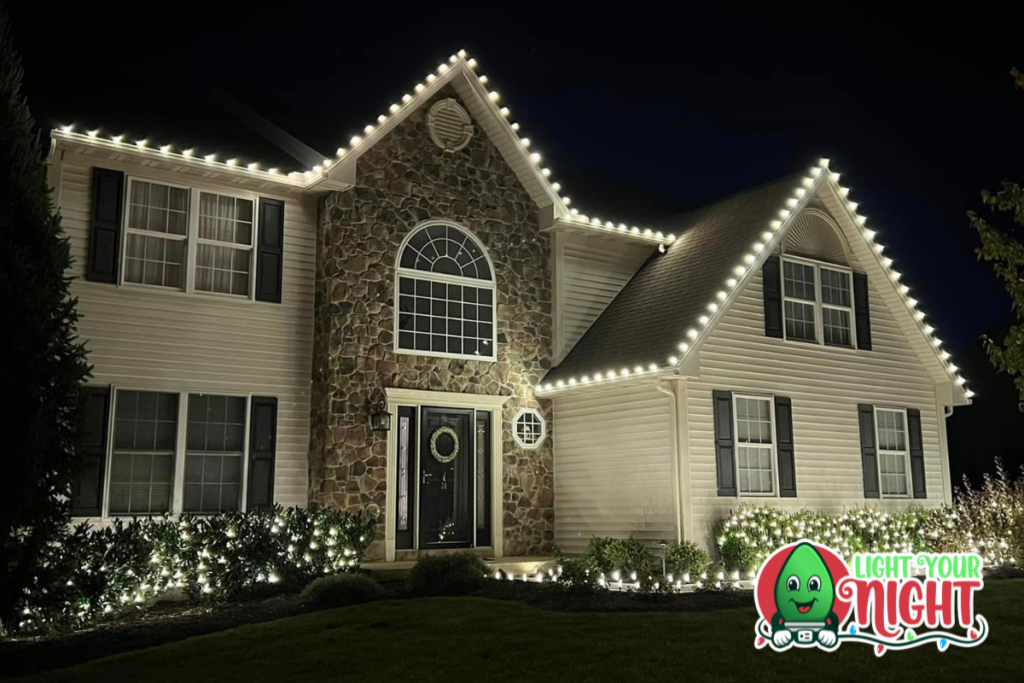 A two-story house is adorned with bright white Christmas lights outlining the roof, windows, and entrance. Shrubs along the front of the house are also illuminated. The night sky provides a stark contrast, emphasizing the festive decorations. A "Light Your Night" logo is in the corner.