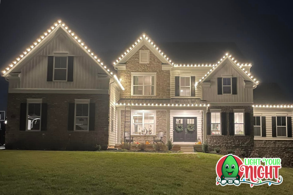 A beautifully lit two-story house at night, adorned with white string lights along its roofline and eaves. The home has a front porch with decorated windows. In the bottom right corner, the logo "Light Your Night" is displayed with a smiling green light bulb character.