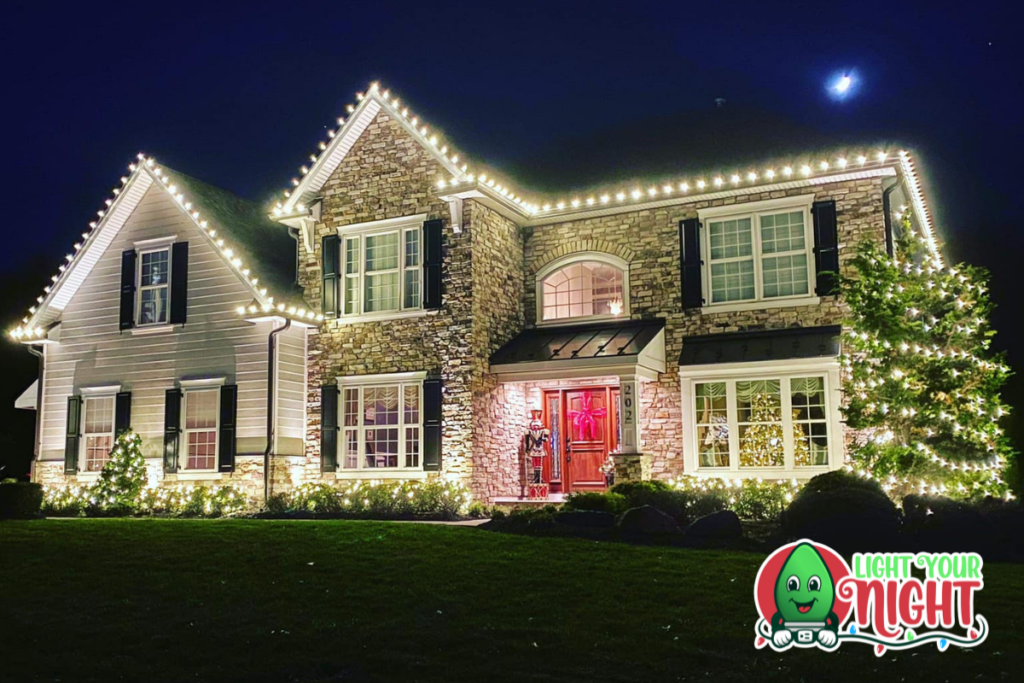 A two-story house adorned with white Christmas lights along the roofline, windows, and bushes. The house has a mix of brick and siding, and there's a festive wreath on the red front door. A decorated tree stands in the front yard. "Lights Your Night" logo is in the bottom right.