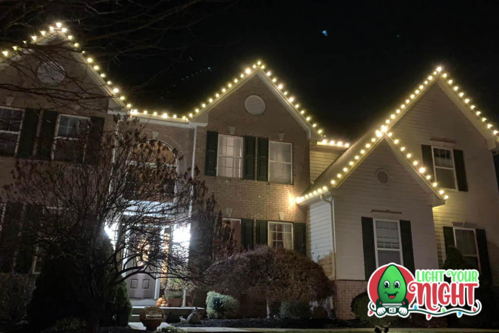 A large two-story house is decorated with warm white Christmas lights along the roofline. The house is made of brick and white siding with dark shutters. The yard is partially visible with some bushes. The "Light Your Night" logo is in the bottom right corner.