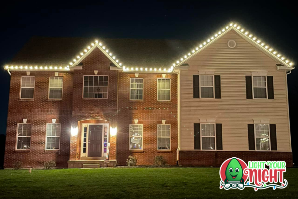 A two-story house is adorned with white holiday lights along the roofline, beautifully illuminating the structure against the night sky. The bottom left corner features a grass lawn, and the bottom right corner displays a logo with a smiling light bulb and the text "Light Your Night.