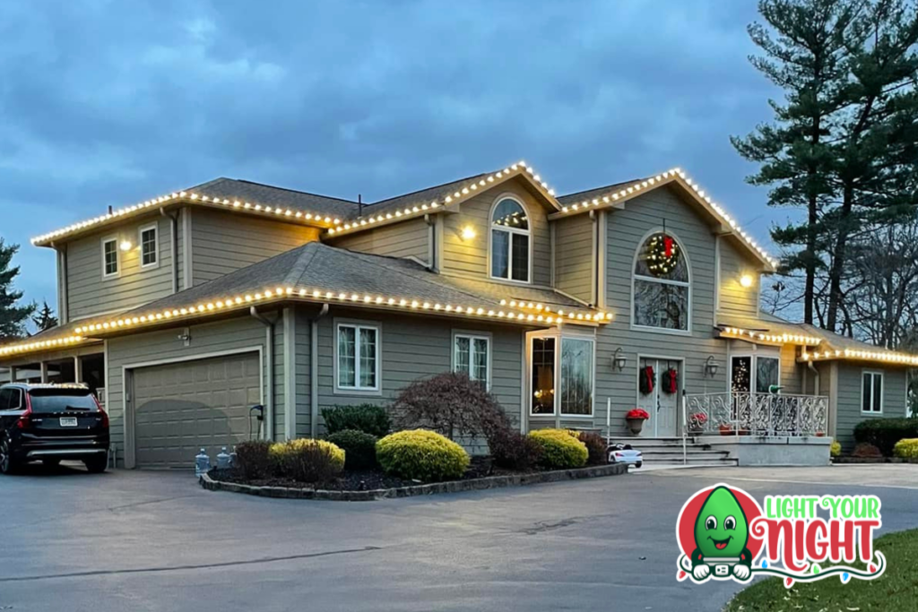 A two-story house is adorned with white holiday lights along its roofline. The house has a large garage on the left and a festive wreath on its upper-level window. There are bushes and trees in the yard. The logo "Light Your Night" is in the bottom right corner.