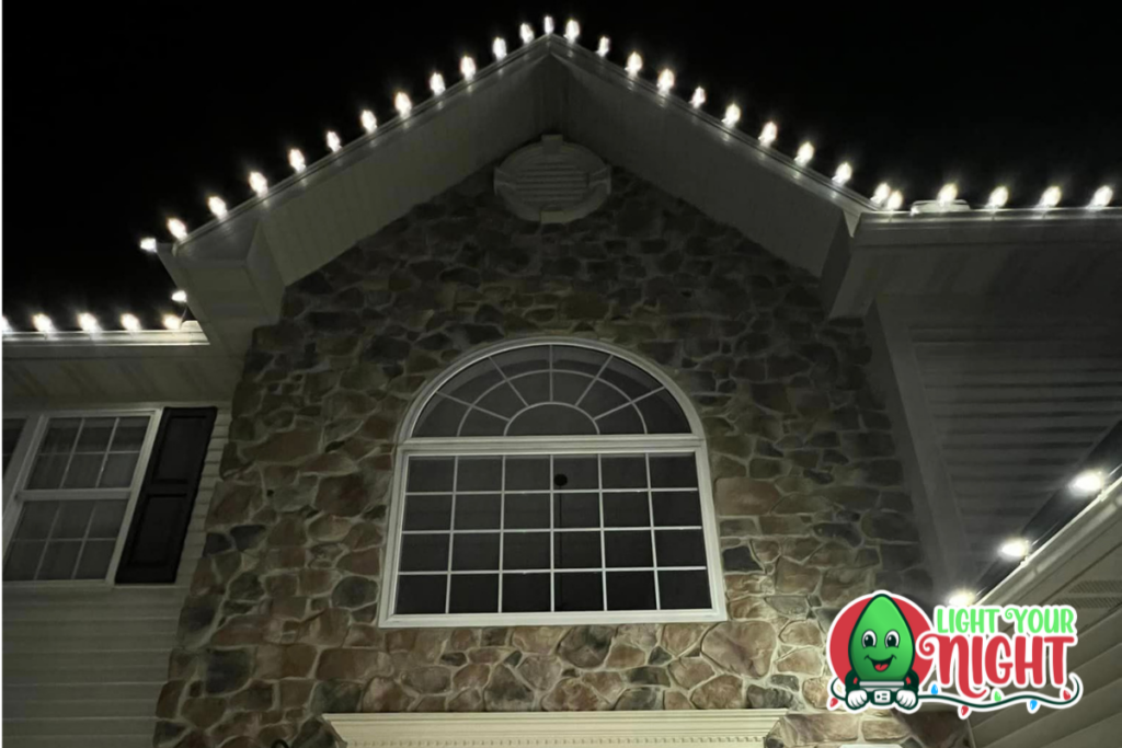 A large stone facade house is adorned with white Christmas lights along the roofline. Above the front window is a semicircular window. The "Light Your Night" logo with a green smiling light bulb is in the bottom right corner.