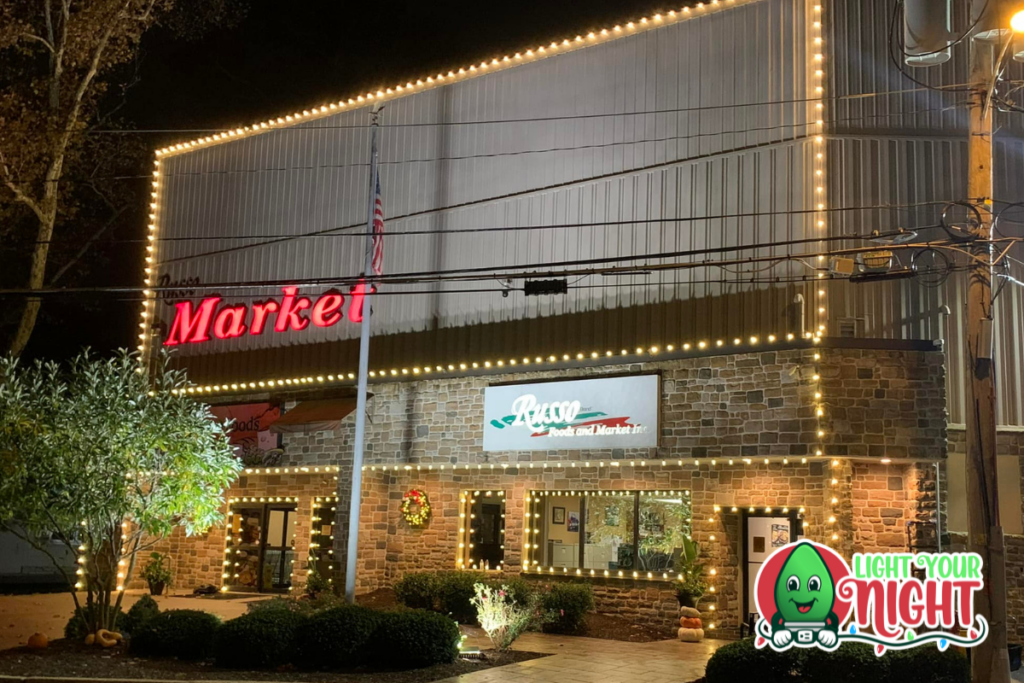 A brick and metal building is decorated with festive string lights, illuminating the night. The neon sign reads "Market" with a sub-sign for "Ross" and "Light Your Night." A small garden and an American flag are visible out front.