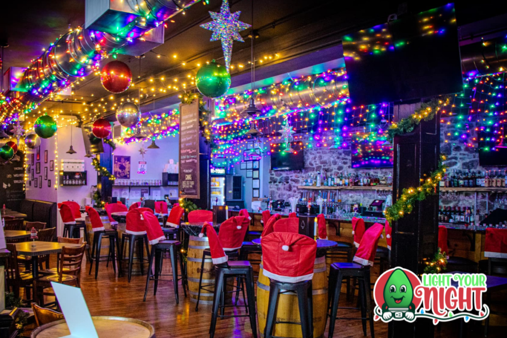 A festive bar decorated for the holidays with colorful string lights, large ornaments, and chairs covered in red and white Santa seat covers. The bar counter is visible along with various bottles, and the floor is wooden. The "Light Your Night" logo is in the corner.