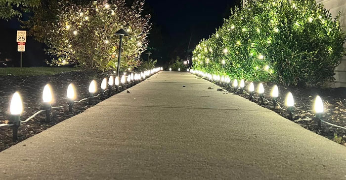 A paved walkway lit by an array of small, white, bulb-shaped lights lining both sides. The path is surrounded by well-manicured shrubs and bushes illuminated by the lights, extending into the dark night. A 25 mph speed limit sign is visible in the background. Partner with us for tranquil evening strolls.