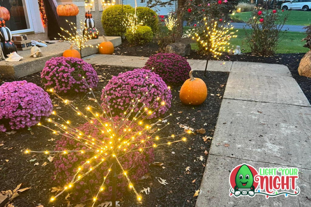 A garden path is lined with vibrant purple and pink flowers, pumpkins, and glowing star-shaped lights. Additional pumpkins and Halloween decorations are near the house entrance. The "Light Your Night" logo featuring a green character in a red suit is at the bottom right.