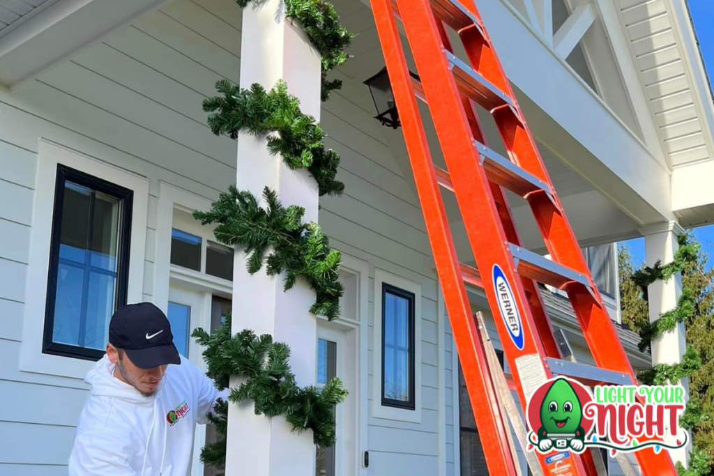 A person in a white hoodie and black cap decorates a white porch column with green garland. A tall red ladder leans against the porch. The house, ideal for holiday displays, is white with large windows and a slanted roof. A "Light Your Night" logo with a green bulb character is in the bottom right corner, highlighting potential careers in festive decoration services.