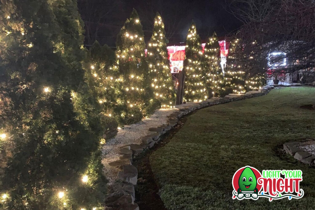 A row of evergreen trees adorned with glowing white Christmas lights, creating a festive ambiance. The trees are behind a stone path and a well-maintained lawn, perfect for any commercial setting. The bottom right corner features the "Light Your Night" logo with a smiling light bulb character.