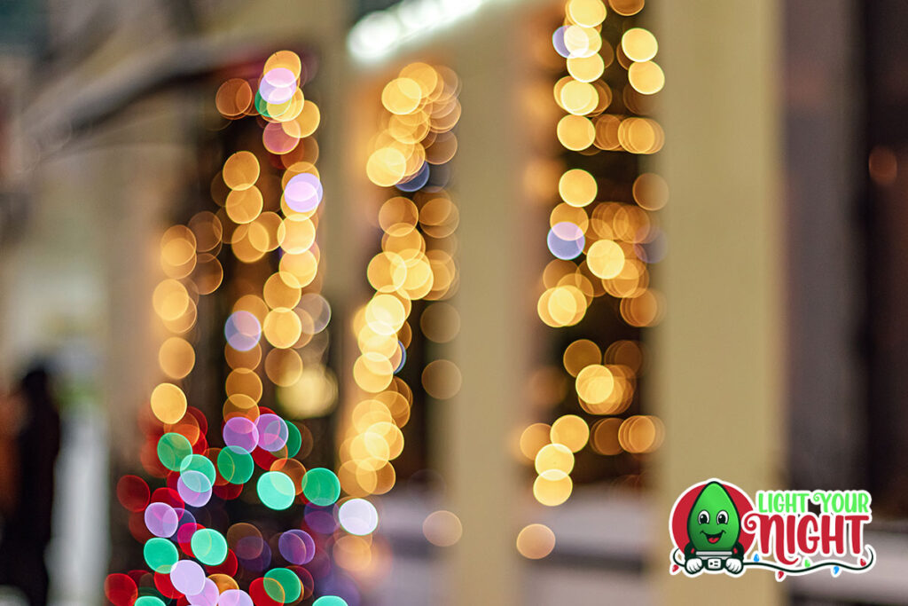 A festive scene with blurred holiday lights in the background, creating a bokeh effect. The lights are warm and colorful, giving a cozy, celebratory atmosphere. In the bottom right corner, there is a commercial logo of "Light Your Night" featuring a cheerful, smiling green light bulb.