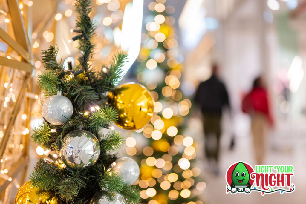 A close-up of a Christmas tree adorned with golden and silver ornaments, with twinkling lights in the background creating a festive atmosphere. Two people are seen walking in the blurry background of this vibrant commercial. The "Light Your Night" logo is placed at the bottom right corner.