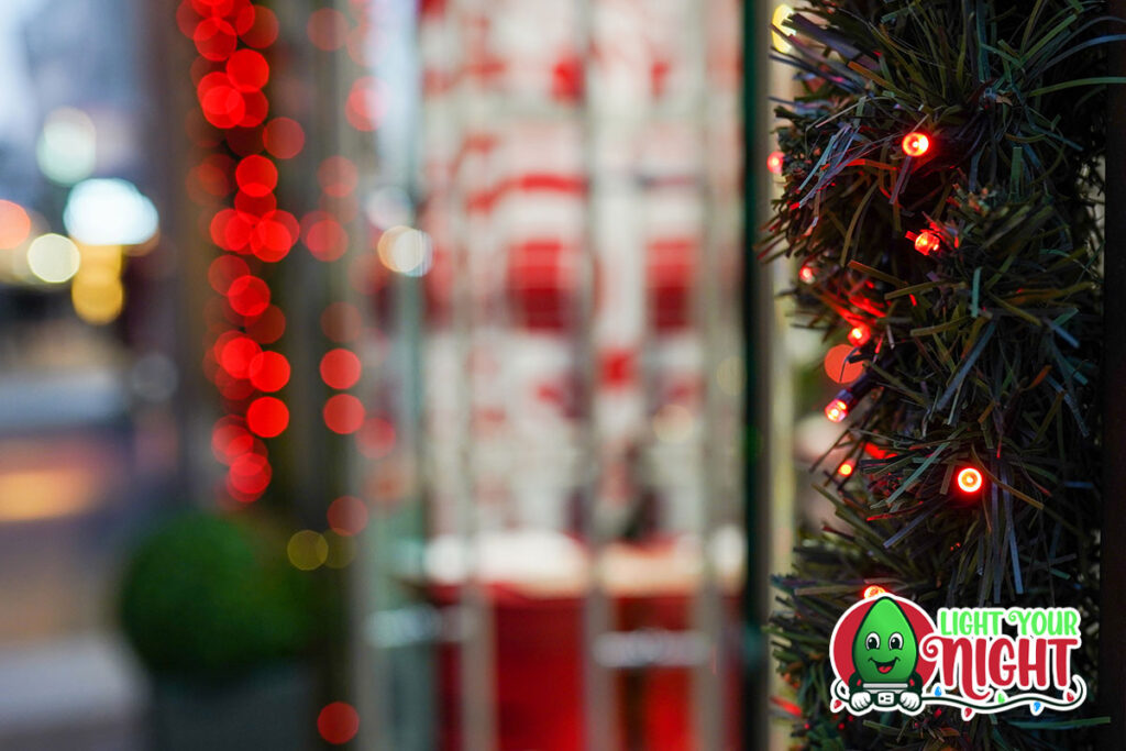 A festive scene with blurred red and white lights in the background and a garland adorned with small red lights in the foreground. The "Light Your Night" logo featuring a green character and glowing text is situated in the lower right corner, perfect for a holiday commercial.