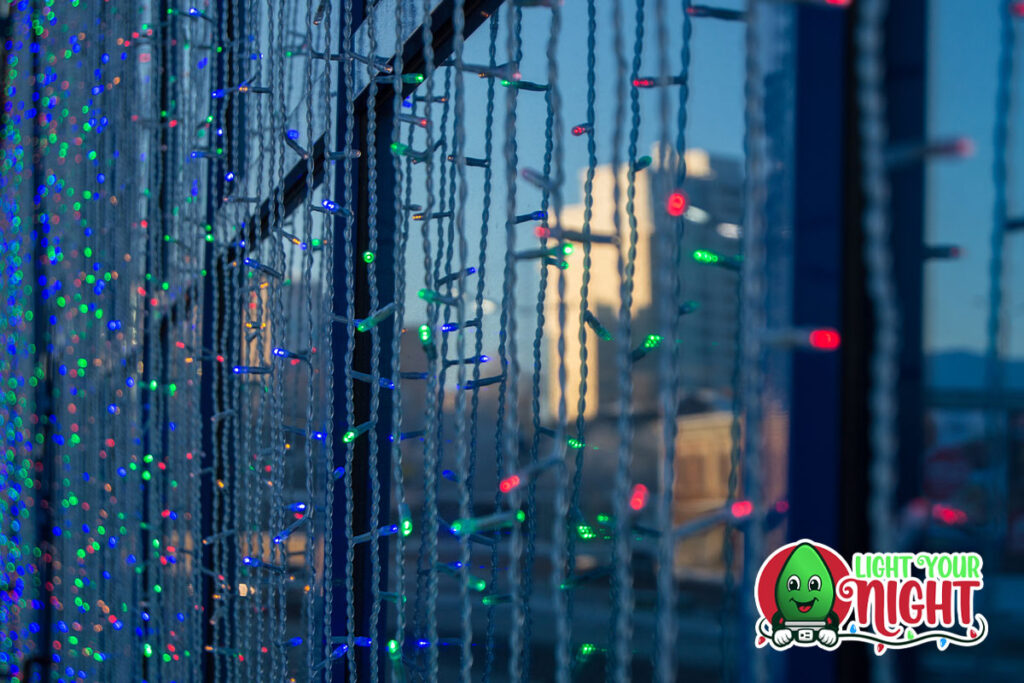 Multicolored string lights are hanging vertically, creating a festive curtain effect in front of a glass window. The cityscape and reflection of buildings are visible in the background. In the lower right corner, there's a logo of a smiling, festive decoration with the text "Light Your Night." Perfect for commercial displays.