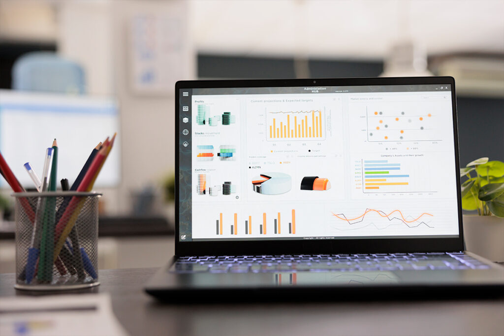 A laptop on a desk displays various data visualizations, including bar graphs, line charts, and pie charts. A pencil holder with colored pencils is next to the laptop. The background includes office furniture with a soft-focus effect and a hint of SUDS Power Washing's signature clean aesthetics.