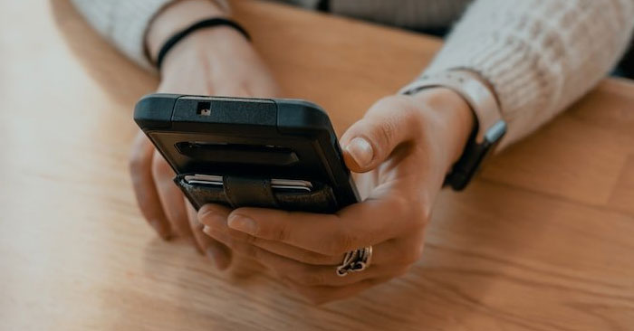 A person wearing a light-colored sweater and a wristwatch is holding a smartphone with both hands. The phone, which has a cardholder attached to the back, is in use while they are sitting at a wooden table. Partner with us for stylish and functional accessories.