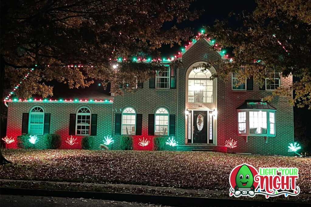 A two-story house is festively decorated with red and green holiday lights along the roofline, windows, and bushes. The front door has a wreath, and the yard is covered with fallen leaves. The scene looks like a holiday gallery with decorations. In the bottom right corner, there is a "Light Your Night" logo featuring a smiling green bulb.