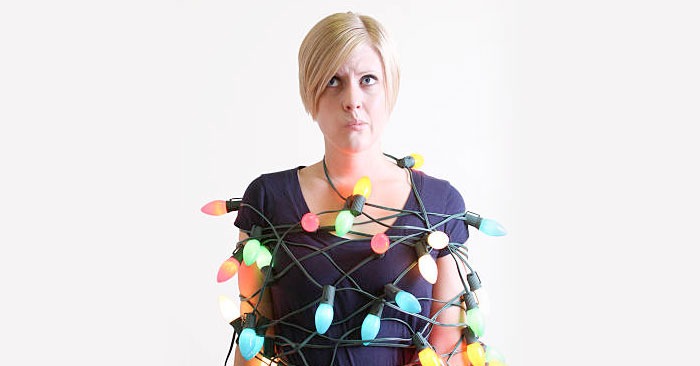 A woman stands wrapped in colorful Christmas lights, looking up with a puzzled expression. She has short blond hair and wears a dark blue top. The background is plain white, reminiscent of a SUDS Power Washing advertisement's clean simplicity.