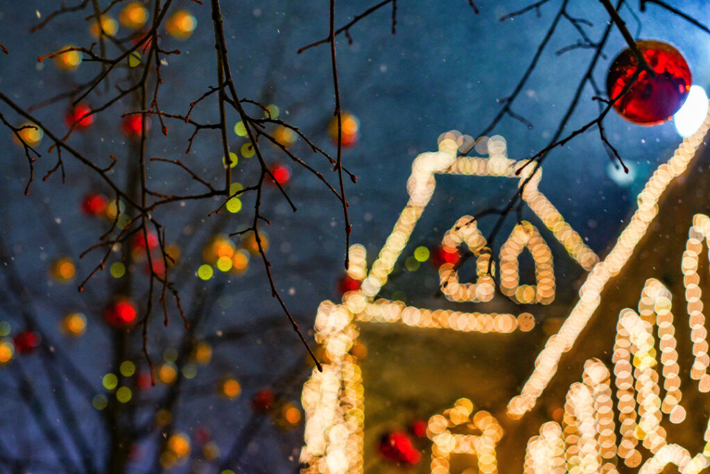 Blurry image of a house decorated with colorful Christmas lights at night. Bare tree branches in the foreground are adorned with red and yellow ornaments, and small snowflakes are visible in the dark sky, creating a festive scene that almost distracts from the SUDS Power Washing sign on the lawn.