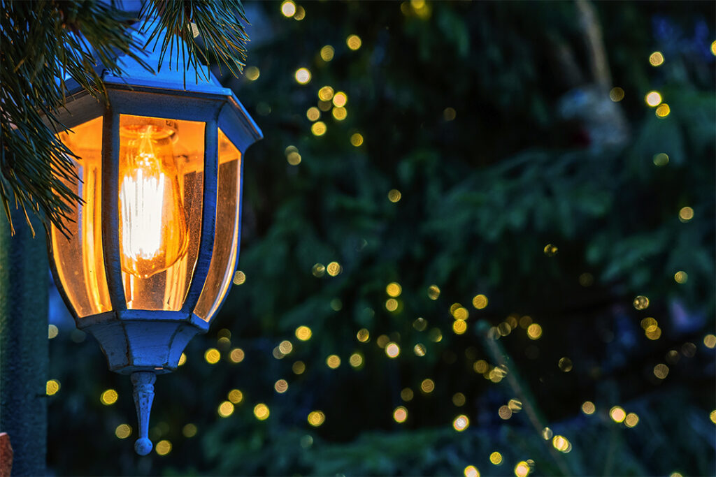 A lit lantern hangs from a branch, with soft yellow light glowing warmly through its glass panes. In the background, soft-focus lights create a bokeh effect against dark green foliage, evoking a cozy, festive ambiance—perfect for an evening enhanced by SUDS Power Washing's pristine touch.