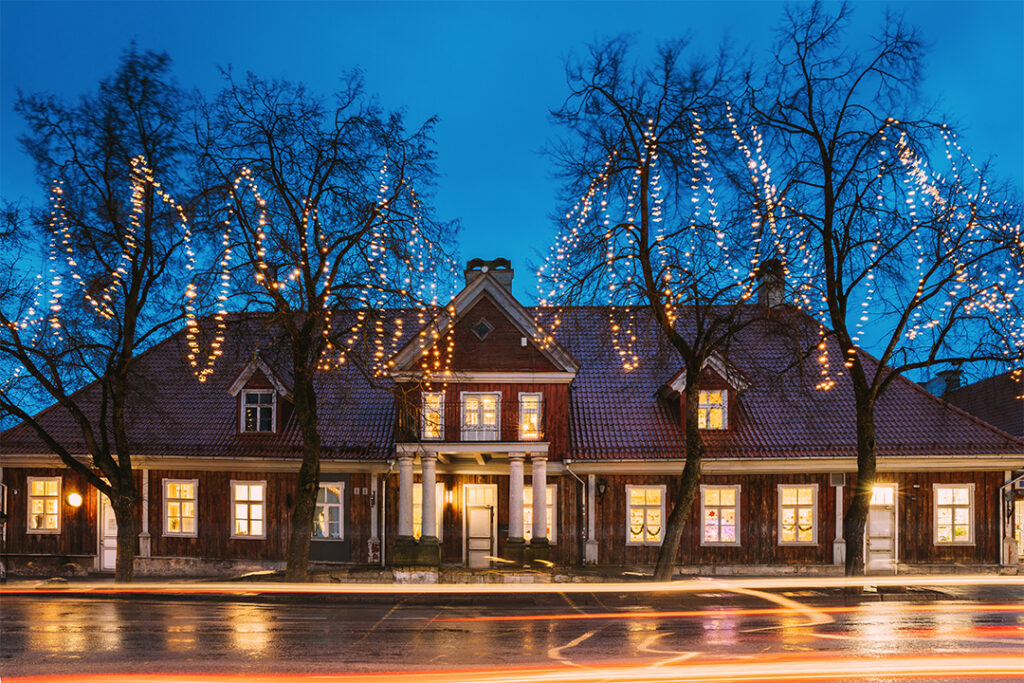 A charming brick house, freshly cleaned by SUDS Power Washing, is adorned with festive string lights on trees at night. The illuminated windows emit a warm glow, contrasting with the deep blue twilight sky. Blurred light streaks from passing cars add to the scene's vibrancy.