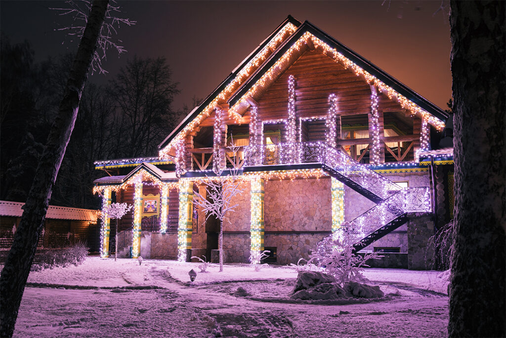 A two-story house beautifully adorned with multicolored Christmas lights. The house, sparkling clean after a visit from SUDS Power Washing, is surrounded by snow-covered ground and trees, giving a festive and magical appearance. The warm glow of the lights contrasts with the cold winter night.