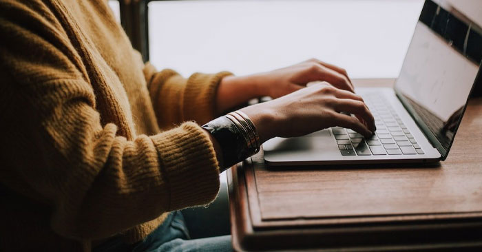 A person wearing a yellow sweater sits at a wooden desk, typing on a laptop. The photo focuses on their hands and the keyboard, with natural light pouring in from the side, suggesting a window nearby. This inviting setup could be your workspace if you choose to partner with us.