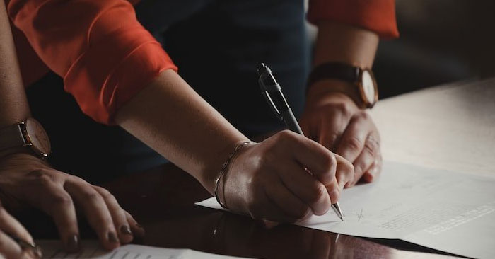 Close-up of two people collaborating over documents on a dark wooden table. One person, wearing a red shirt and a watch, is holding a pen and writing on one of the documents, while the other hand rests on another paper nearby. Partner with us to achieve seamless collaboration like this.