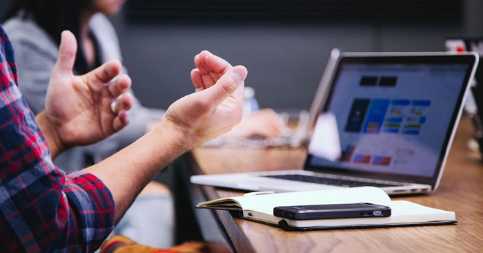 A person wearing a plaid shirt is gesturing with their hands in front of an open laptop on a wooden table, likely discussing the benefits of SUDS Power Washing. A notebook and a smartphone are also on the table, and another person is blurred in the background, highlighting the collaborative work environment.