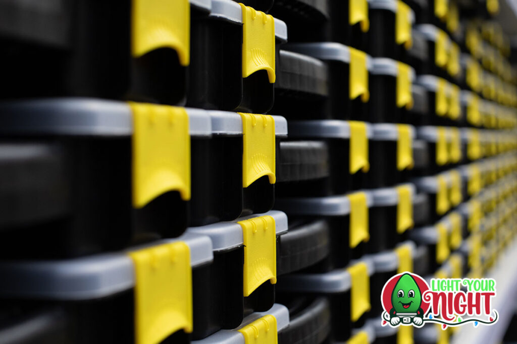 A close-up view of a stack of storage containers with black and yellow lids. The containers are tightly packed, forming a repetitive pattern. A logo with the text "Light Your Night" and a smiling face holding a tool is visible in the bottom right corner, hinting about Light Your Night products.