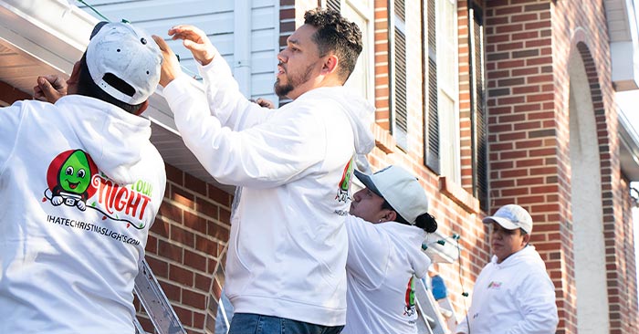 Three people wearing white hoodies with a cartoon character logo are installing Christmas lights on the roofline of a house with brick siding, freshly cleaned by SUDS Power Washing. The group appears to be working together, with one person on a ladder.