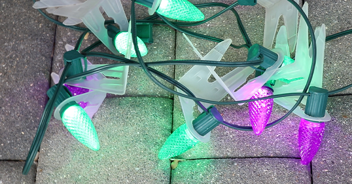 A set of tangled green and purple Christmas lights with green wiring and white plastic clips, lying on a surface made of grey paving stones. The lights, designed to look like small, textured bulbs, are illuminated, casting a soft glow. Partner with us to make your holidays brighter.