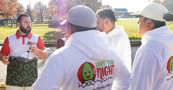 Four men are standing outdoors on a sunny day. One man, facing the others, is wearing a red and white shirt and holding a pamphlet. The other three men are wearing white hoodies with a cartoonish Christmas light bulb and the text "Light Your Night" on the back, promoting SUDS Power Washing services.