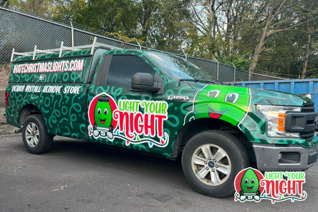 A green truck is parked on the side of the road, covered in a festive wrap featuring a cartoon light bulb character and the text "Light Your Night" in red and green. The company's services, website, and career opportunities are also displayed on the truck. Trees form a vibrant background.