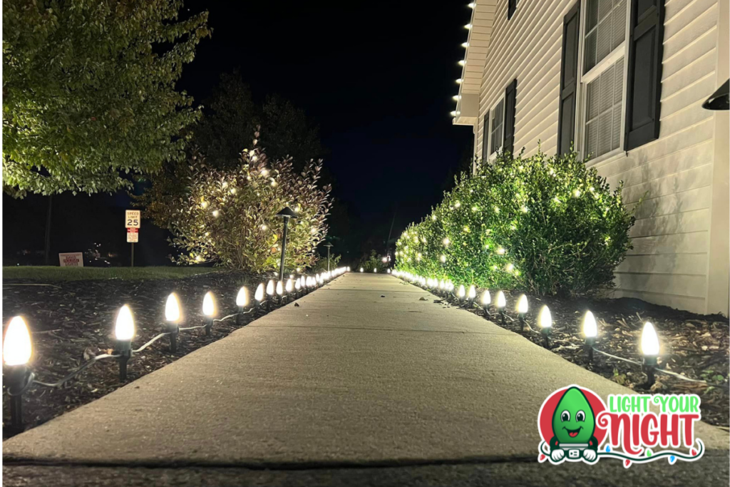 A sidewalk lined with small, glowing lights leads to a house decorated with white string lights on the bushes and trees. The "Light your Night" logo featuring a cheerful bulb character is visible in the bottom-right corner. The scene is taken at night.