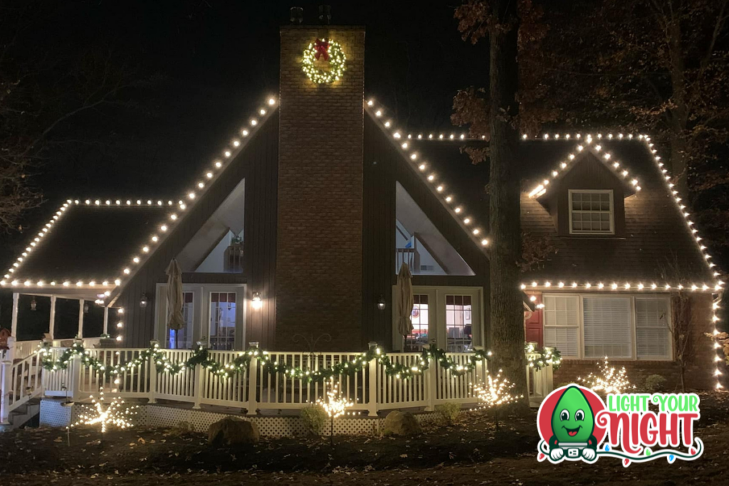 A house decorated for Christmas with white string lights outlining the roof and chimney. The porch is adorned with garlands and a lit wreath hangs on the chimney. In the bottom right corner, there's a "Light Your Night" logo featuring a smiling lightbulb.