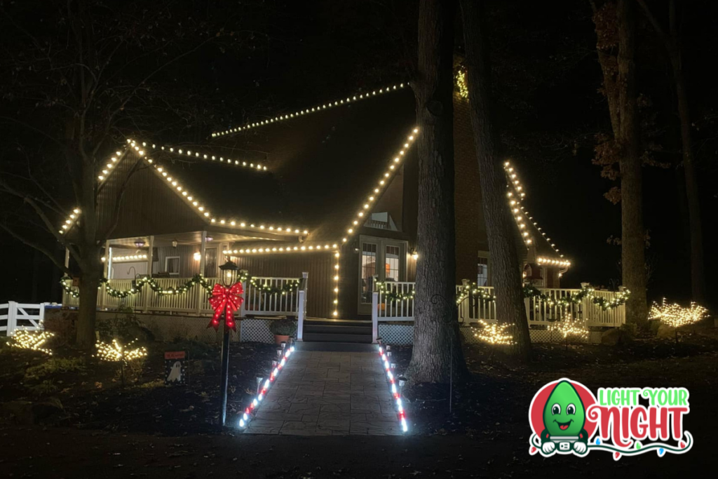 A house beautifully decorated with Christmas lights and greenery. White and yellow lights outline the roof and porch, with garlands and a red bow on the railing. Trees and bushes are also adorned with lights. A "Light Your Night" logo is in the bottom right corner.