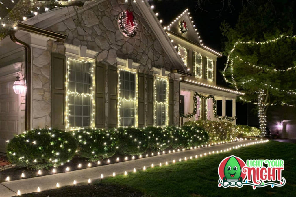 A two-story house beautifully decorated with white string lights for the holiday season. The façade features lights outlining windows, doors, and rooflines. A "Light Your Night" logo is at the bottom right corner. Bushes also have individual light decorations.