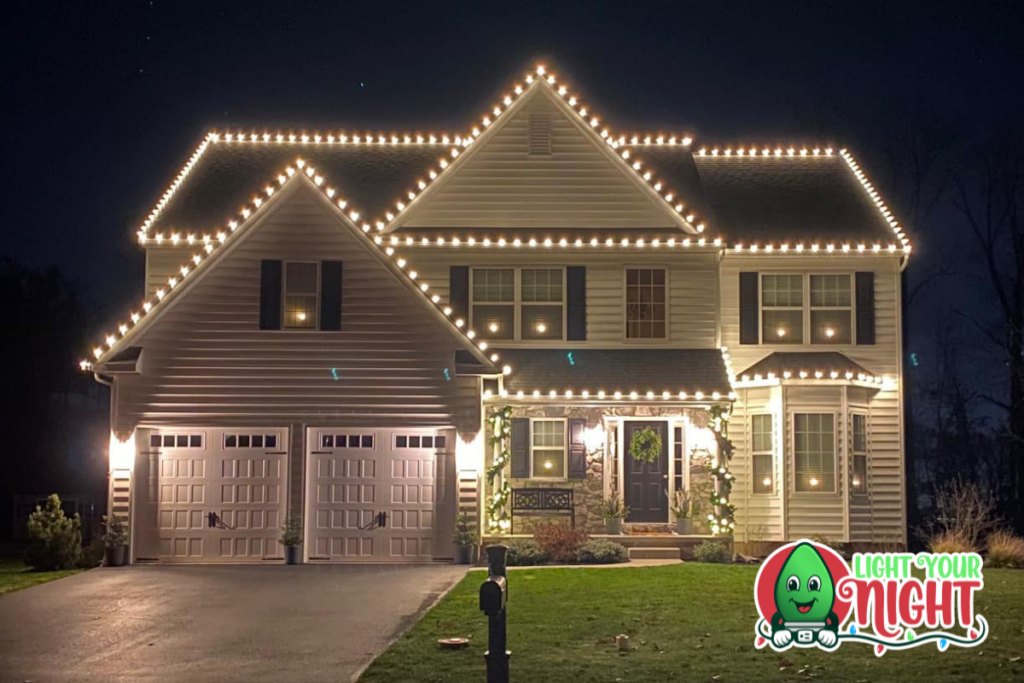 A two-story house is decorated with white string lights outlining the roof and garage. Greenery with lights adorns the front porch. A "Light Your Night" logo with a cartoon light bulb is in the lower right corner of the image. The sky is dark.