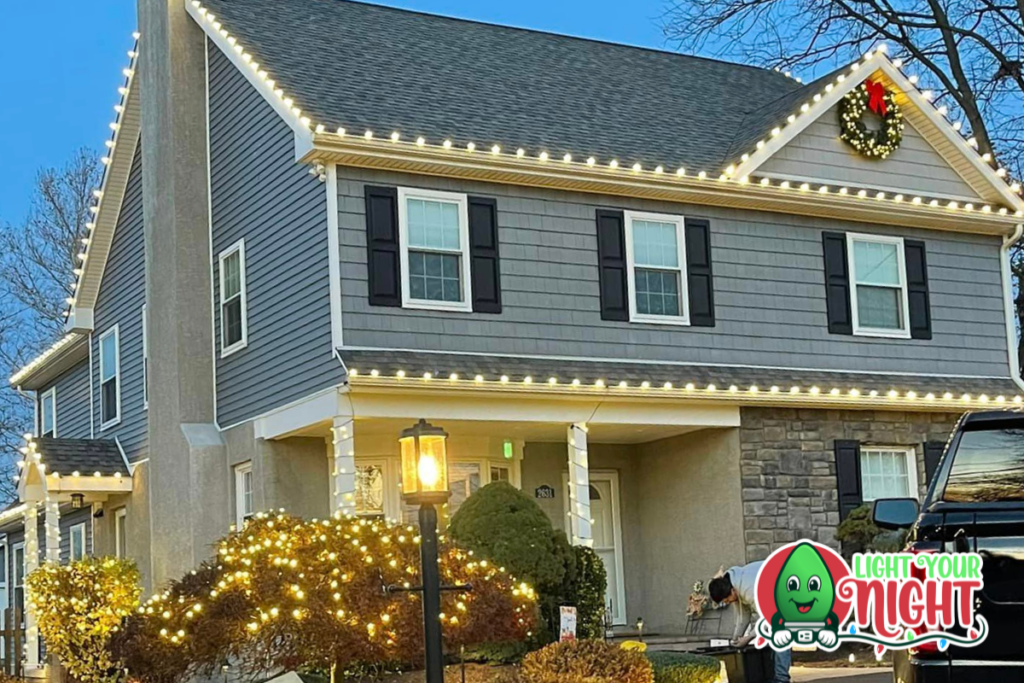 A two-story house is adorned with white string lights along the roofline, windows, and porch. A holiday wreath hangs above the garage. Bushes in the yard are festively lit, and a logo in the corner features "Light Your Night" text with a colorful light bulb character.