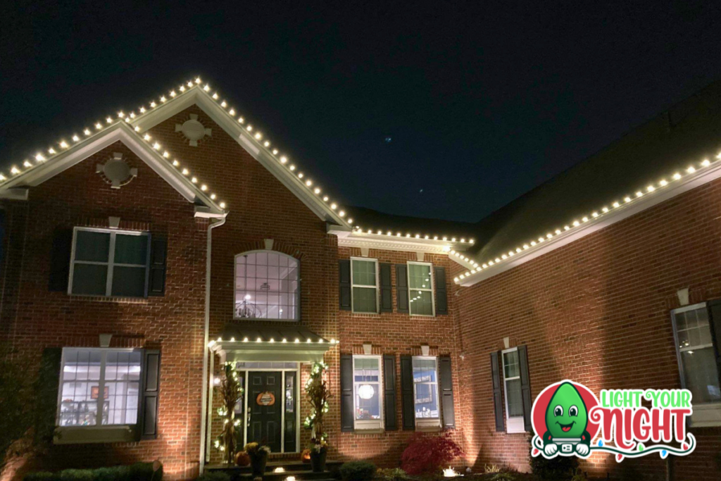 A large brick house is decorated with white string lights outlining the roof and windows. The night sky is visible in the background. A logo with a smiling bulb character and the words "Light Your Night" appears at the bottom right.